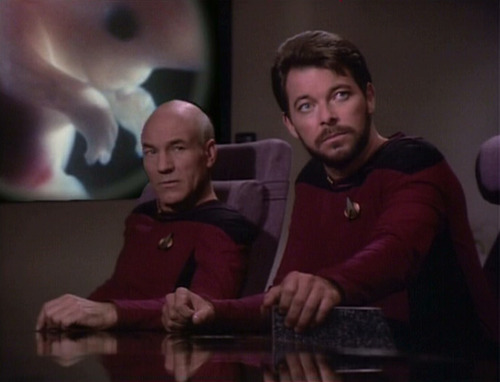 Riker and Picard sit at conference table in front of image of Troi's fetus