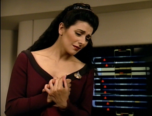 Troi clasps Riker's hand to her chest as he lies in his Sickbay bed