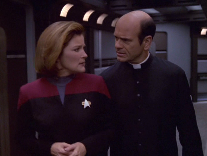 The Doctor dressed as his Priest character walks and talks with Janeway in the corridor