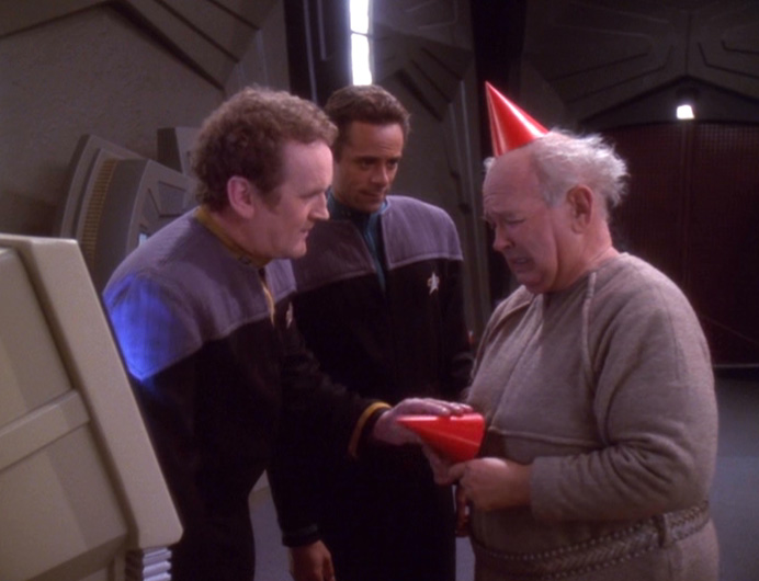 Patrick, an older man in a red paper birthday hat, tries to give a hat to Miles