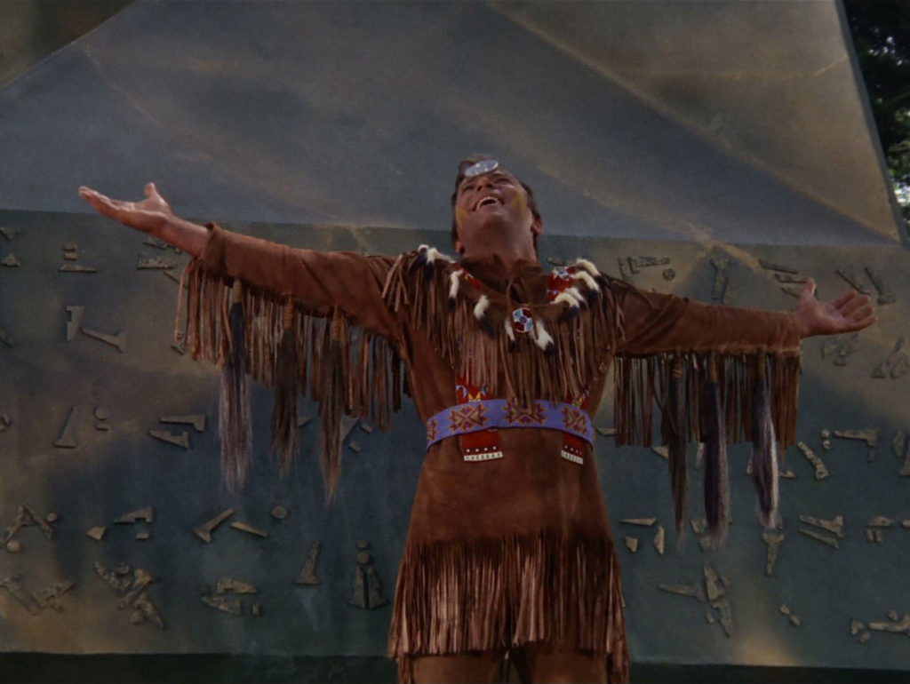 Kirk as Kirok, in buckskins and a headband spreads his arms wide