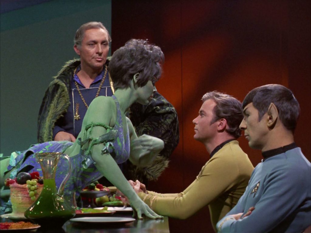 Marta dances, lying across the dinner table in front of Kirk and Spock