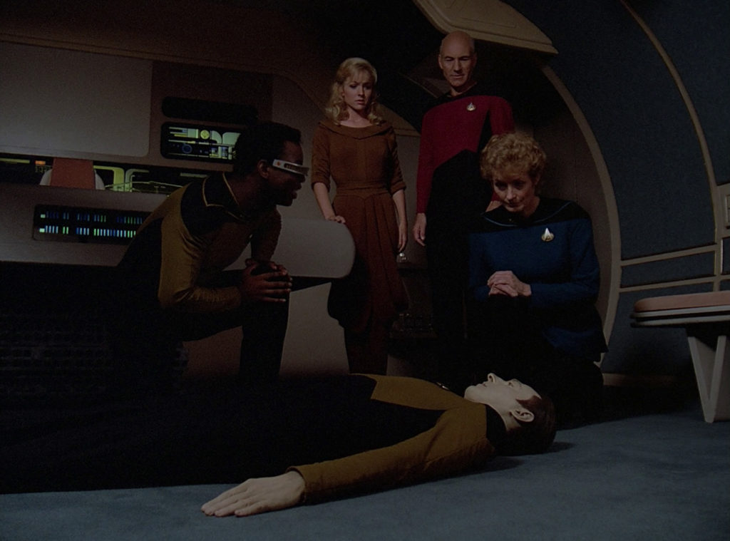 Data lying on the ground while Pulaski and La Forge kneel over him, Kareen and Picard look on